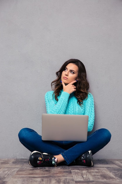 Portret van een peinzende vrouw zittend op de vloer met laptop op grijze muur