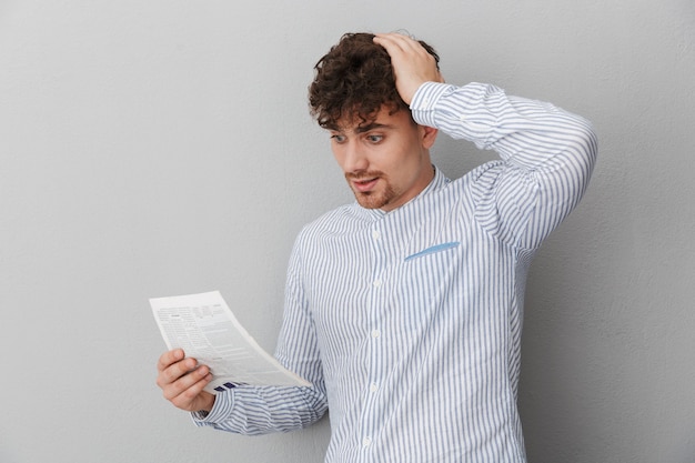 Portret van een perplexe jongeman gekleed in een shirt die zijn hoofd grijpt terwijl hij een krant of tijdschrift vasthoudt en leest, geïsoleerd over een grijze muur