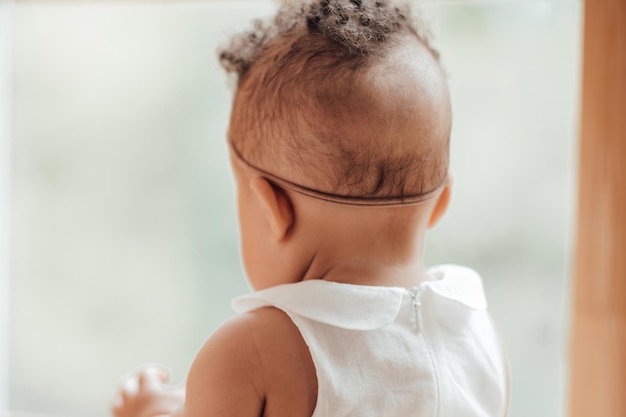 Portret van een peutermeisje van gemengd ras in een witte jurk met babyspeelgoed dat bij het raam zit en wegkijkt