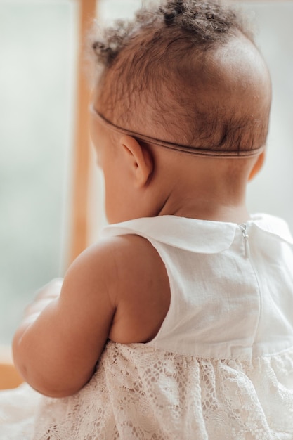 Portret van een peutermeisje van gemengd ras in een witte jurk met babyspeelgoed dat bij het raam zit en wegkijkt