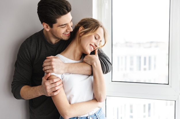 Portret van een positief stel dat lacht en samen knuffelt terwijl ze in de buurt van een groot raam in een lichte kamer thuis staan