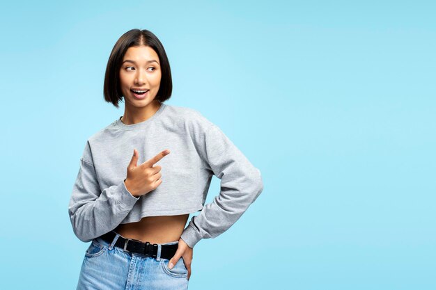 Foto portret van een positieve aantrekkelijke aziatische vrouw in casual kleding die met de vinger naar de kopierruimte wijst en wegkijkt en geïsoleerd op een blauwe achtergrond staat.