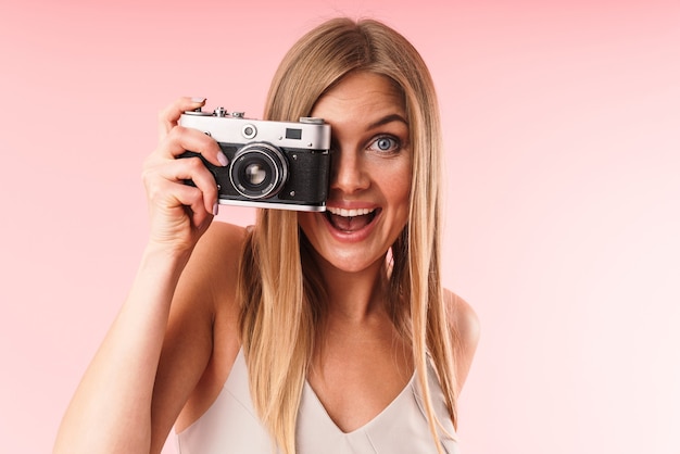 Portret van een positieve blonde vrouw die een jurk draagt die lacht terwijl ze een portret maakt op een retrocamera die over een roze muur in de studio wordt geïsoleerd