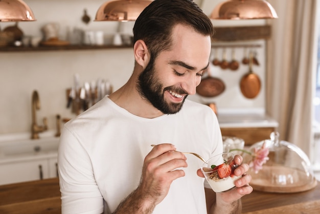 Portret van een positieve brunette man die panna cotta-dessert met theelepel eet terwijl hij thuis ontbijt in een stijlvolle keuken