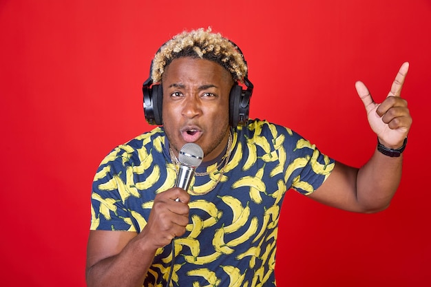 Portret van een positieve genietende mooie Afrikaanse man met een microfoon en koptelefoon op zijn hoofd, luisterend naar muziek die een lied zingt dat op een rode achtergrond wordt geïsoleerd