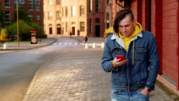 Portret van een positieve jonge man met dreadlocks die mobiele telefoon doorbladert op moderne stadsstraat