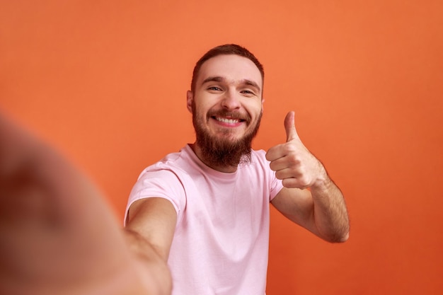 Portret van een positieve knappe bebaarde man die selfie maakt of streamt, duim omhoog POV laat zien, oogpunt van foto, roze T-shirt draagt. Indoor studio opname geïsoleerd op een oranje achtergrond.