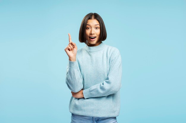 Foto portret van een positieve mooie aziatische vrouw in casual kleding met een vinger omhoog die een goed idee heeft en naar de camera kijkt, geïsoleerd op een blauwe achtergrond.