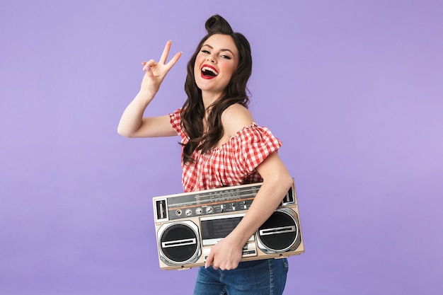 Portret van een positieve pin-up vrouw in Amerikaanse stijl die zich verheugt terwijl ze een oude vintage boombox vasthoudt die over een violette muur wordt geïsoleerd