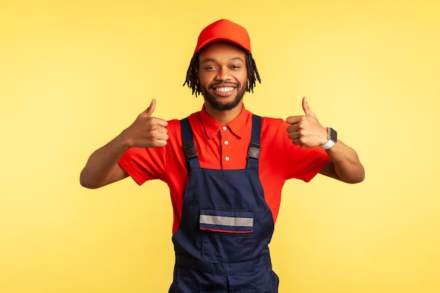 Portret van een positieve reparateur met een overall die duimen omhoog laat zien terwijl hij naar de camera kijkt, adviseert de dienst huishouden onderhoud Indoor studio shot geïsoleerd op gele achtergrond