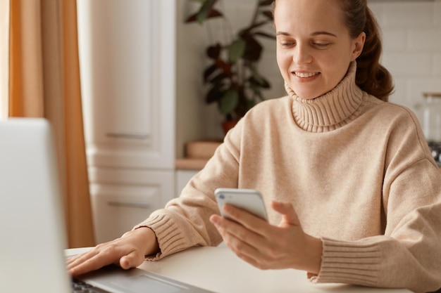 Portret van een positieve schattige vrouw die een beige casual stijl trui draagt met een paardenstaartkapsel die aan tafel voor een laptopscherm zit en mobiele telefoon gebruikt voor het controleren van e-mails.