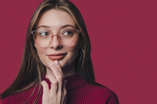 Portret van een positieve vrouw met een bril die nadenkt over een perfecte weekendaanrakingskin met handen geïsoleerd over karmozijnrode kleurachtergrond