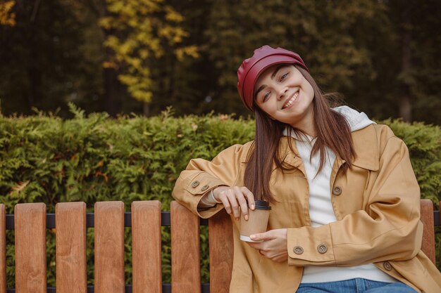 Portret van een positieve vrouw met een kopje koffie om te gaan rusten in het herfstpark en naar de camera te kijken