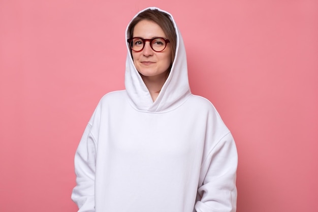Portret van een positieve zelfverzekerde vrouw in witte hoodie en bril op roze achtergrond in studio