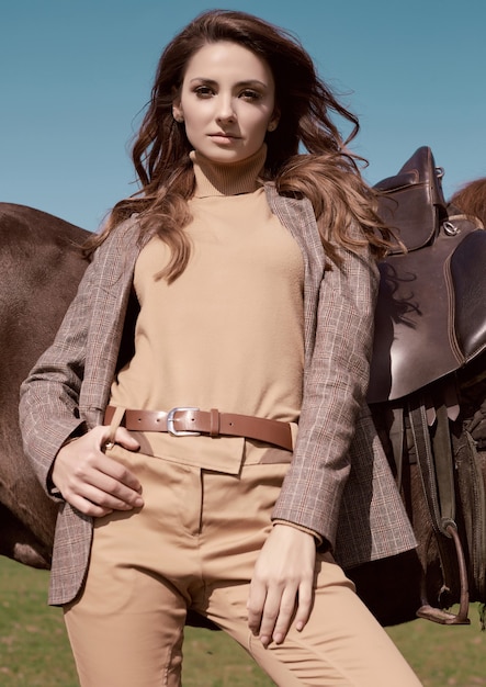 Portret van een prachtige brunette vrouw in een elegant geruit bruin jasje poseren met een paard op landlandschap