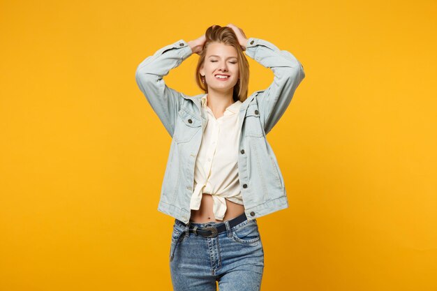 Portret van een prachtige jonge vrouw in denim vrijetijdskleding die staat, de handen op het hoofd zet, de ogen gesloten houdt geïsoleerd op de geeloranje muurachtergrond. Mensen levensstijl concept. Bespotten kopie ruimte.