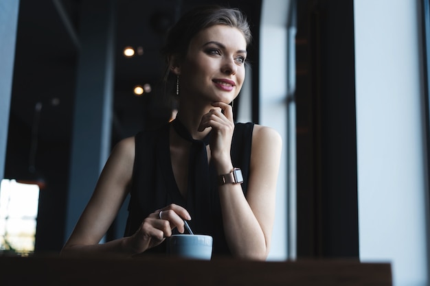 Portret van een prachtige vrouw die thee of koffie drinkt en met een glimlach uit het raam van de coffeeshop kijkt terwijl ze geniet van haar vrije tijd, leuke zakenvrouwenlunch in een modern café tijdens haar werkpauze.