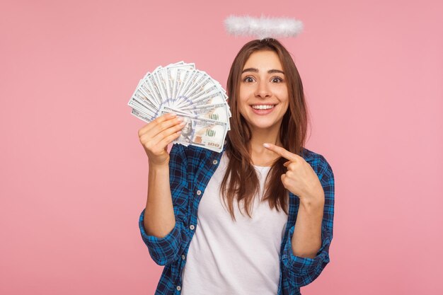 Portret van een rijk engelachtig meisje, loterijwinnaar met halo boven het hoofd die geld wijst en gelukkig glimlacht, dollarbiljetten laat zien, genietend van het rijke paradijsleven. studio-opname geïsoleerd op roze achtergrond