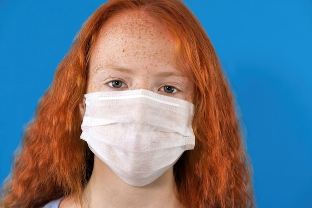 portret van een roodharig meisje in een wit medisch masker met blauwe ogen en sproeten op blauw