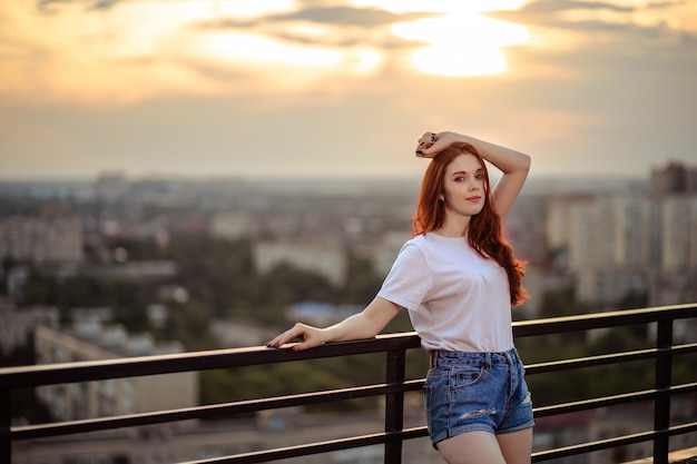 Portret van een roodharig meisje op het dak van een hoog huis bij zonsondergang met uitzicht op de stad