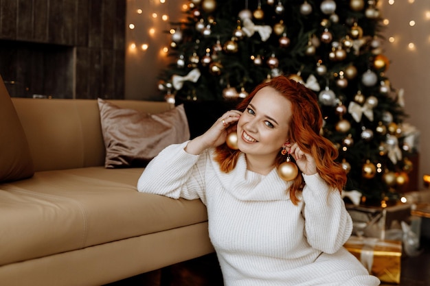 Portret van een roodharige vrouw in een witte trui glimlachend tegen de achtergrond van een kerstboom