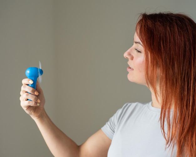 Portret van een roodharige vrouw met een zakventilator in haar handen Profiel van een meisje verfrist in de hitte door een wind die wordt geblazen door een draadloos elektrisch apparaat Geniet van de koele luchtstroom van de airconditioner