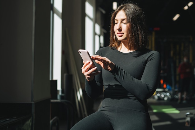 Portret van een rustende jonge brunette vrouw in sportactieve kleding met behulp van een mobiele telefoon in een fitnessclub