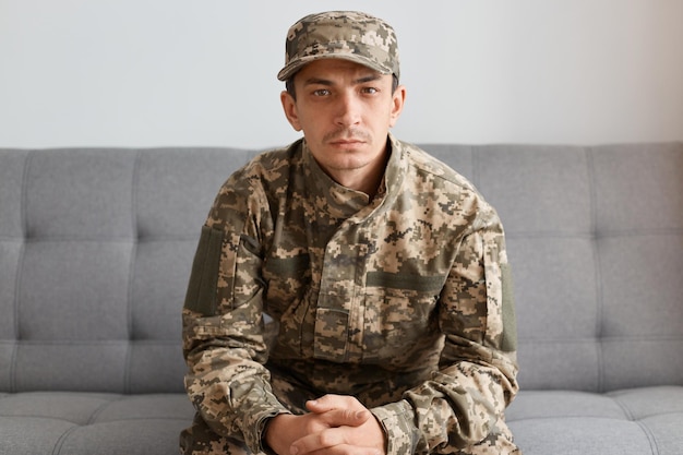 Portret van een rustige, serieuze blanke militaire man met een camouflage-uniform en een pet die op een bank zit en naar de camera kijkt met een depressieve gezichtsuitdrukking