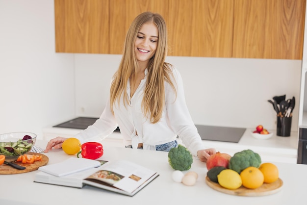 Portret van een schattig blond meisje glimlachend en lezend een receptenboek in de keuken op tafel tussen ingrediënten om te koken