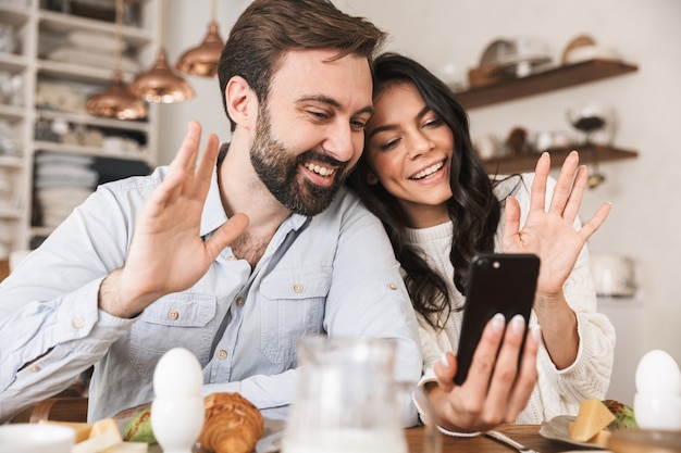 Portret van een schattig Europees stel man en vrouw die smartphone gebruiken terwijl ze thuis ontbijten in de keuken