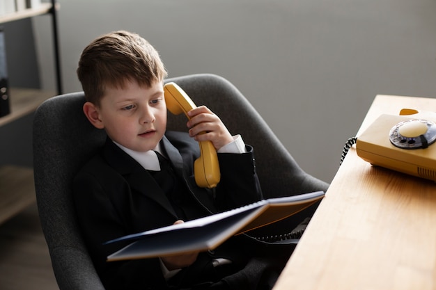 Foto portret van een schattig kind in een pak dat aan het bureau zit en een roterende telefoon gebruikt