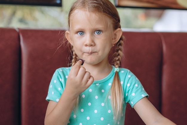 Portret van een schattig kindmeisje met blond haar en blauwe ogen in een mooi blauw polka dot T-shirt dat naar de camera kijkt en iets interessants proeft.