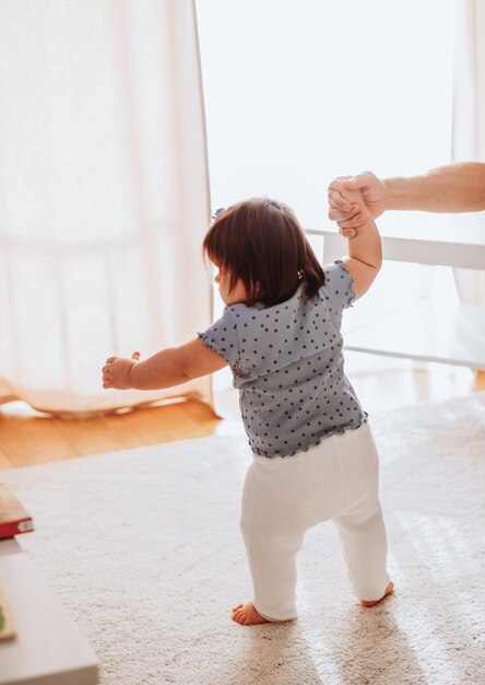 Portret van een schattig klein babymeisje dat thuis leert lopen terwijl ze vaders handen vasthoudt, kleine peuter-baby