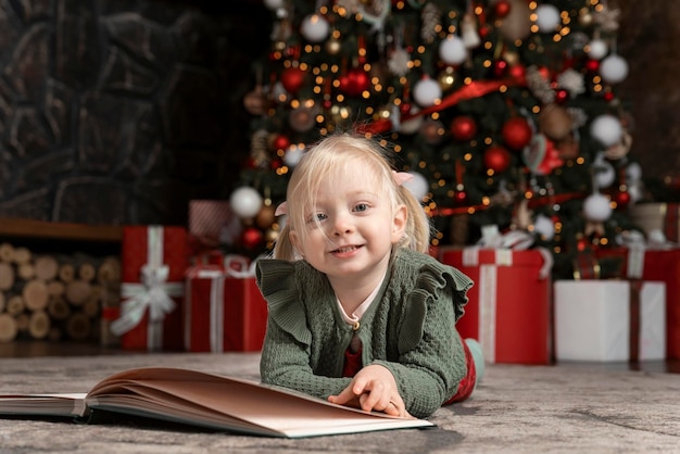 Portret van een schattig klein blond meisje dat op de vloer ligt met een groot boek onder de kerstboom Kinderen en nieuwjaarsstemming