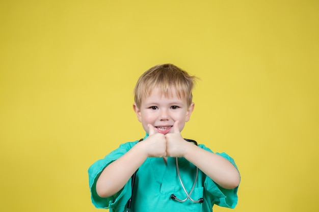 Portret van een schattig klein kaukasisch kind gekleed in groene dokters met duim omhoog gebaar op gele achtergrond