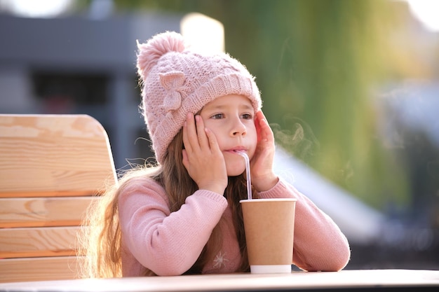 Portret van een schattig klein kindmeisje met een roze hoed die alleen zit in een straatcafé en thee drinkt uit een papieren bekertje