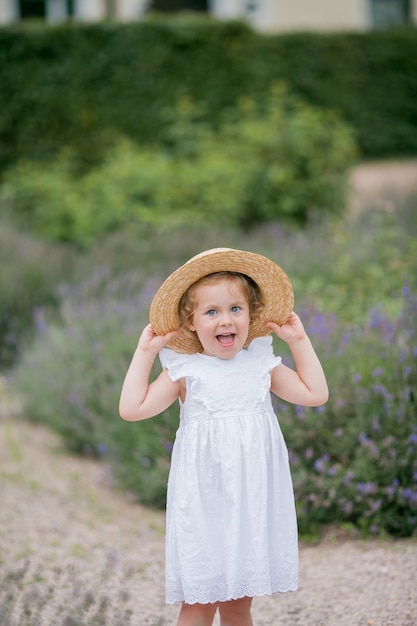 Portret van een schattig klein krullend blauwogig meisje van 3 jaar oud in een rieten hoed en witte zomerjurk in de buurt van bloeiend lavendel gelukkig kind in de natuur