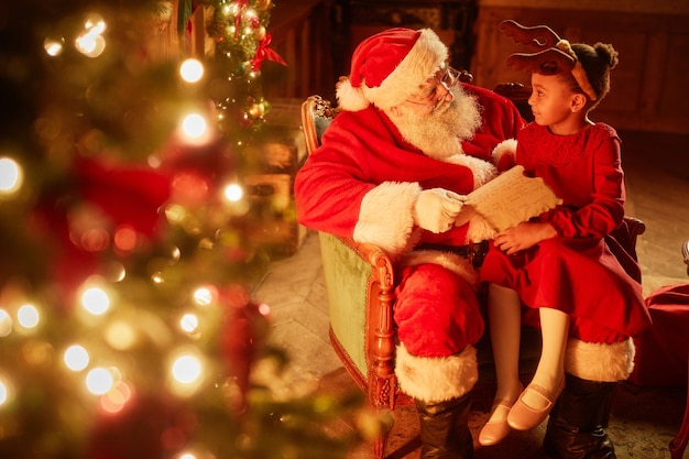 Portret van een schattig klein meisje dat in de schoot van de kerstman zit bij de kerstboom in een magische set kopieerruimte