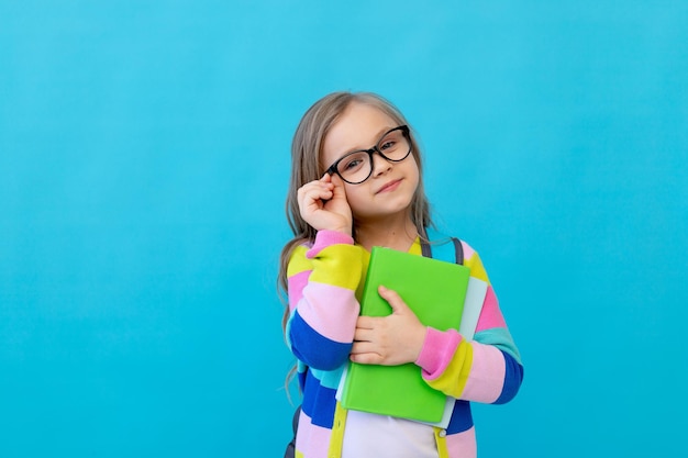 Portret van een schattig klein meisje met een bril in een gestreept jasje met notebooks en een rugzak past haar bril aan als een leraar Concept van onderwijs Fotostudio blauwe achtergrond plaats voor tekst