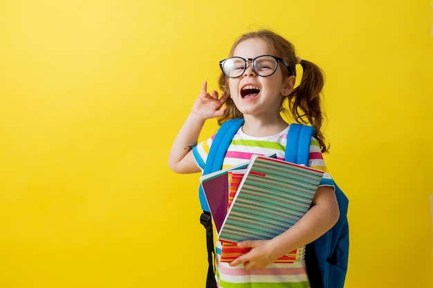 Foto portret van een schattig klein meisje met een bril in een gestreept t-shirt met notitieboekjes en schoolboeken in haar handen en een rugzak. onderwijsconcept. fotostudio, gele achtergrond, ruimte voor tekst.