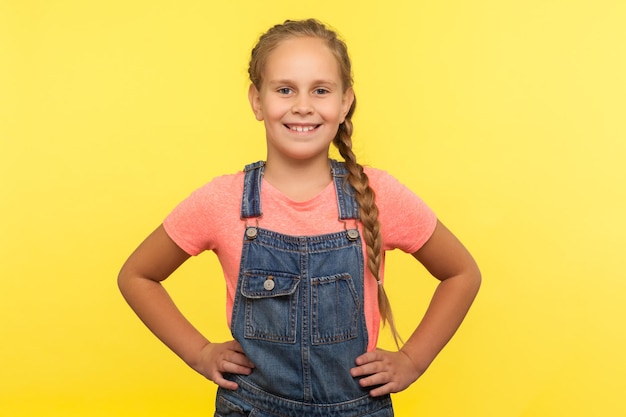 Foto portret van een schattig klein meisje met een vlecht in een denim overall die handen op de heupen houdt en glimlacht naar de camera, een gelukkig kind dat vertrouwensoptimisme uitdrukt. indoor studio-opname geïsoleerd op gele achtergrond