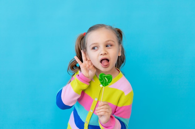 Portret van een schattig klein meisje met lolly's in een gestreept jasje. het kind eet en likt een grote lolly. het concept van snoep en snoep. fotostudio, blauwe achtergrond, plaats voor tekst