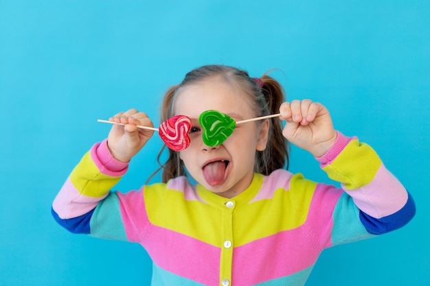 portret van een schattig klein meisje met lolly's in een gestreepte jas die haar ogen ermee bedekt een groot snoepje op een stok het concept van snoep en snoepjes blauwe achtergrondruimte voor tekst