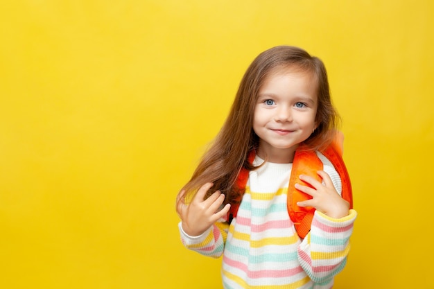 Portret van een schattig klein schoolmeisje met een rugzak in een trui op een geel gekleurde achtergrond