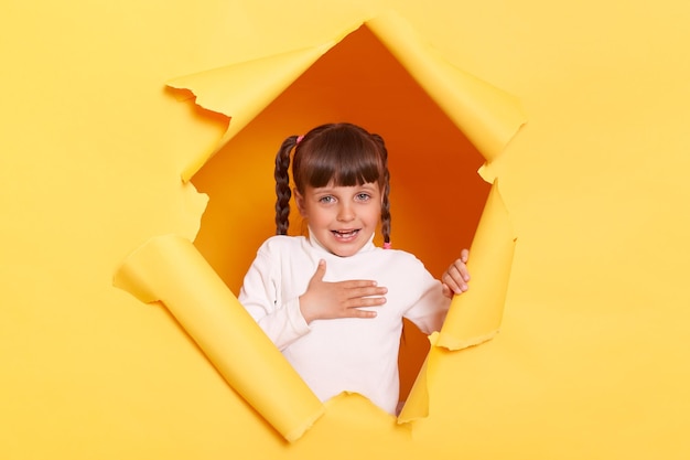 Portret van een schattig schattig klein meisje met vlechten met een witte coltrui die poseert in een gescheurd gat van een gele papieren muur houdt de hand op de borst en is blij dat ze is gekozen