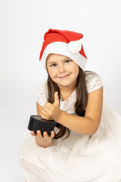 Portret van een schattig zesjarig meisje met een kerstmuts die een slimme muziekluidspreker vasthoudt en duimen opgeeft...