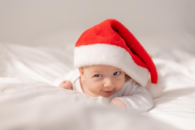 Portret van een schattige baby in de hoed van de kerstman liggend op zijn buik in bed. charmant kind met blauwe ogen. wintervakantie, grappige momenten, kinderkerst. ruimte voor tekst. Hoge kwaliteit foto