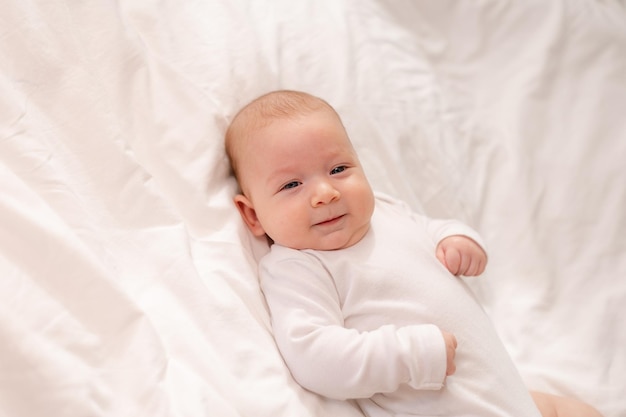 Portret van een schattige baby met blauwe ogen in een witte bodysuit die op de rug op wit beddengoed ligt