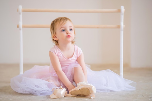 Portret van een schattige babyballerina met blauwe ogen in roze, zittend in de buurt van de balletbarre die pointe-schoenen aantrekt