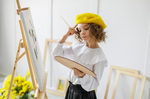 Portret van een schattige, bedachtzame kleine meisjekunstenaar in een gele baret die een foto in de studio schildert terwijl hij een kleurenpalet vasthoudt